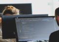 Two people work at computers with code visible on the screen, set in an office with a focused atmosphere. One wears a striped shirt; the other has glasses.