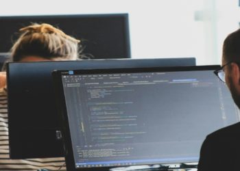 Two people work at computers with code visible on the screen, set in an office with a focused atmosphere. One wears a striped shirt; the other has glasses.