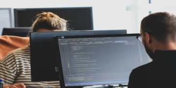 Two people work at computers with code visible on the screen, set in an office with a focused atmosphere. One wears a striped shirt; the other has glasses.