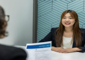 Woman wearing a navy blazer and white blouse sits confidently at a table during an interview. She is smiling, facing the interviewer, creating a positive and engaging atmosphere. The background features a geometric patterned wall.