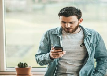 Man in a denim shirt sits on a windowsill, focused on his phone. A small potted cactus is beside him. The scene is calm and casual.