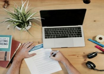 Person writes on paper at a wooden desk with a laptop, a book on marketing, a potted plant, headphones, and stationery, conveying a focused work setting.