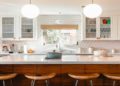 Modern kitchen with a large marble island, wooden stools, and hexagonal tile backsplash. White cabinets and pendant lights create a bright, inviting space.