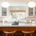 Modern kitchen with a large marble island, wooden stools, and hexagonal tile backsplash. White cabinets and pendant lights create a bright, inviting space.