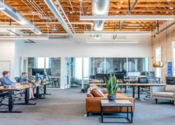 Modern office space with an open layout, featuring wooden beams, desks with people working on computers, a cozy seating area, and several large windows.