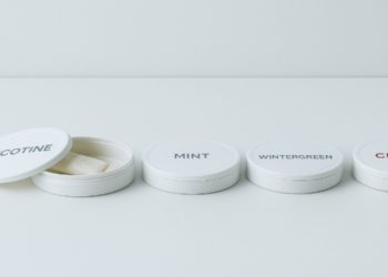 Four white circular tins with labels: Nicotine (lid open showing pouches), Mint, Wintergreen, and Citrus. Minimalist design on a plain surface.
