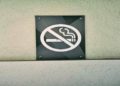 "No Smoking" sign is mounted on a textured white wall. The black sign features a red circle with a diagonal line over a cigarette graphic, conveying prohibition.