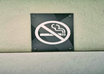 "No Smoking" sign is mounted on a textured white wall. The black sign features a red circle with a diagonal line over a cigarette graphic, conveying prohibition.