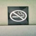 "No Smoking" sign is mounted on a textured white wall. The black sign features a red circle with a diagonal line over a cigarette graphic, conveying prohibition.