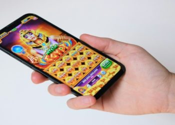 Hand holds a smartphone displaying a colorful slot machine game with an Egyptian theme. The screen shows icons and bright graphics, evoking excitement and fun.
