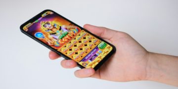 Hand holds a smartphone displaying a colorful slot machine game with an Egyptian theme. The screen shows icons and bright graphics, evoking excitement and fun.