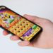 Hand holds a smartphone displaying a colorful slot machine game with an Egyptian theme. The screen shows icons and bright graphics, evoking excitement and fun.