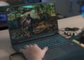 Person playing a video game on a laptop with a colorful backlit keyboard. The screen shows a vibrant outdoor scene. Wires are plugged in, and they wear colorful headphones.