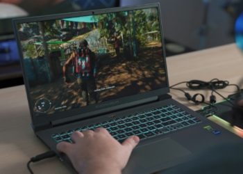 Person playing a video game on a laptop with a colorful backlit keyboard. The screen shows a vibrant outdoor scene. Wires are plugged in, and they wear colorful headphones.