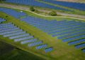 Aerial view of a vast solar farm with rows of blue solar panels on lush green grass. A winding dirt path and a train track run through the scene, evoking progress and sustainability.