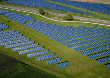 Aerial view of a vast solar farm with rows of blue solar panels on lush green grass. A winding dirt path and a train track run through the scene, evoking progress and sustainability.