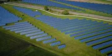Aerial view of a vast solar farm with rows of blue solar panels on lush green grass. A winding dirt path and a train track run through the scene, evoking progress and sustainability.