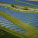 Aerial view of a vast solar farm with rows of blue solar panels on lush green grass. A winding dirt path and a train track run through the scene, evoking progress and sustainability.