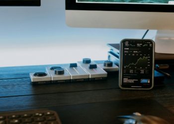 Smartphone displaying stock graphs is on a desk with a keyboard and mouse in front of a computer monitor. The setting feels modern and tech-focused.