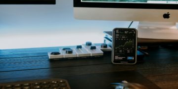 Smartphone displaying stock graphs is on a desk with a keyboard and mouse in front of a computer monitor. The setting feels modern and tech-focused.