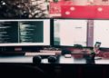 Dual-monitor setup on a desk shows code on one screen and a chat application on the other. Headphones and a smartphone are nearby, suggesting productivity.