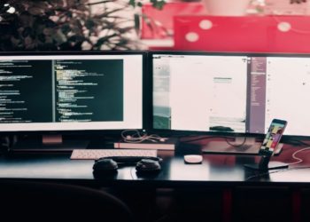 Dual-monitor setup on a desk shows code on one screen and a chat application on the other. Headphones and a smartphone are nearby, suggesting productivity.