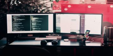Dual-monitor setup on a desk shows code on one screen and a chat application on the other. Headphones and a smartphone are nearby, suggesting productivity.