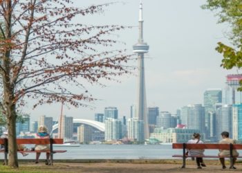 Two people sit on separate benches under leafy trees, facing a calm lake and Toronto Canada's skyline with the CN Tower
