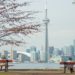 Two people sit on separate benches under leafy trees, facing a calm lake and Toronto Canada's skyline with the CN Tower