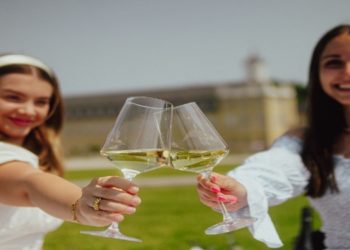 Two women in white and light blue dresses smiling and toasting with wine glasses outdoors in a sunny, green garden setting.