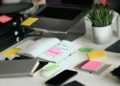 Cluttered desk with an open notebook marked "Done" on a sticky note, surrounded by a laptop, phone, stacked files, various colored sticky notes, and a potted plant. Bright and busy atmosphere.