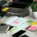 Cluttered desk with an open notebook marked "Done" on a sticky note, surrounded by a laptop, phone, stacked files, various colored sticky notes, and a potted plant. Bright and busy atmosphere.
