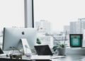 Modern office with a large window view of city buildings. A sleek desk features an iMac, laptop, plant, and tablet, conveying a clean, professional vibe.