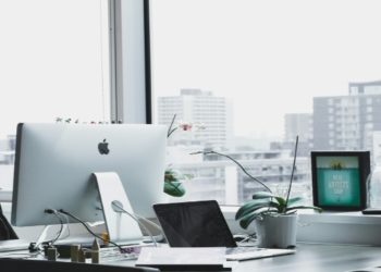 Modern office with a large window view of city buildings. A sleek desk features an iMac, laptop, plant, and tablet, conveying a clean, professional vibe.