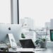 Modern office with a large window view of city buildings. A sleek desk features an iMac, laptop, plant, and tablet, conveying a clean, professional vibe.