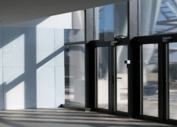 Sunlight streams through large glass doors, casting geometric shadows on a modern building's floor and walls, creating a serene and minimalistic ambiance.