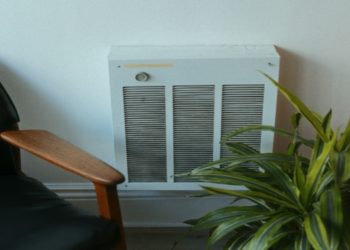 Black chair with wooden armrests sits beside a green leafy plant and a wall-mounted white heater.