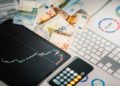 Financial workspace with Euro banknotes, a tablet showing a stock chart, and printed graphs on a desk. A calculator and keyboard are nearby.