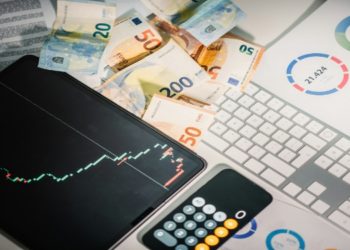 Financial workspace with Euro banknotes, a tablet showing a stock chart, and printed graphs on a desk. A calculator and keyboard are nearby.