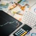Financial workspace with Euro banknotes, a tablet showing a stock chart, and printed graphs on a desk. A calculator and keyboard are nearby.