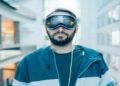 Man in a blue hoodie and Apple Vision Pro futuristic goggles stands in a modern, glass-paneled hallway. The scene conveys a tech-savvy, futuristic vibe.