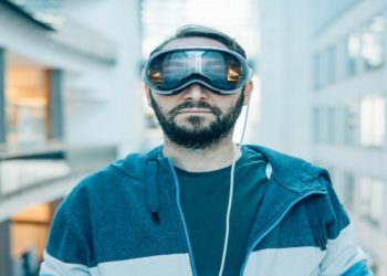 Man in a blue hoodie and Apple Vision Pro futuristic goggles stands in a modern, glass-paneled hallway. The scene conveys a tech-savvy, futuristic vibe.