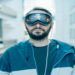 Man in a blue hoodie and Apple Vision Pro futuristic goggles stands in a modern, glass-paneled hallway. The scene conveys a tech-savvy, futuristic vibe.