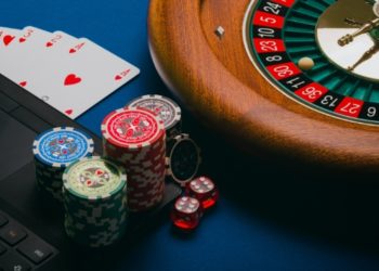 Roulette wheel next to a laptop, poker chips, dice, and playing cards on a blue table, conveying a casino gambling theme. Vibrant and tense atmosphere.