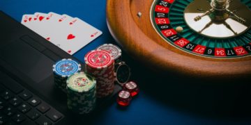 Roulette wheel next to a laptop, poker chips, dice, and playing cards on a blue table, conveying a casino gambling theme. Vibrant and tense atmosphere.