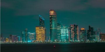 City skyline at night, featuring illuminated skyscrapers with vibrant lights reflecting on the water below. The atmosphere is serene and modern.