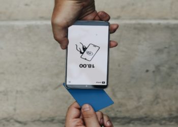 Smartphone held upside down displays "18:00" with a card icon. Below, a hand holds a blue card. The background is a neutral concrete wall.