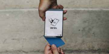 Smartphone held upside down displays "18:00" with a card icon. Below, a hand holds a blue card. The background is a neutral concrete wall.