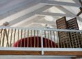 Cozy loft with a wooden railing overlooks a room, featuring a large, curved red sofa and a slatted wooden partition. Exposed beams add rustic charm.