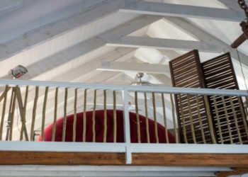 Cozy loft with a wooden railing overlooks a room, featuring a large, curved red sofa and a slatted wooden partition. Exposed beams add rustic charm.